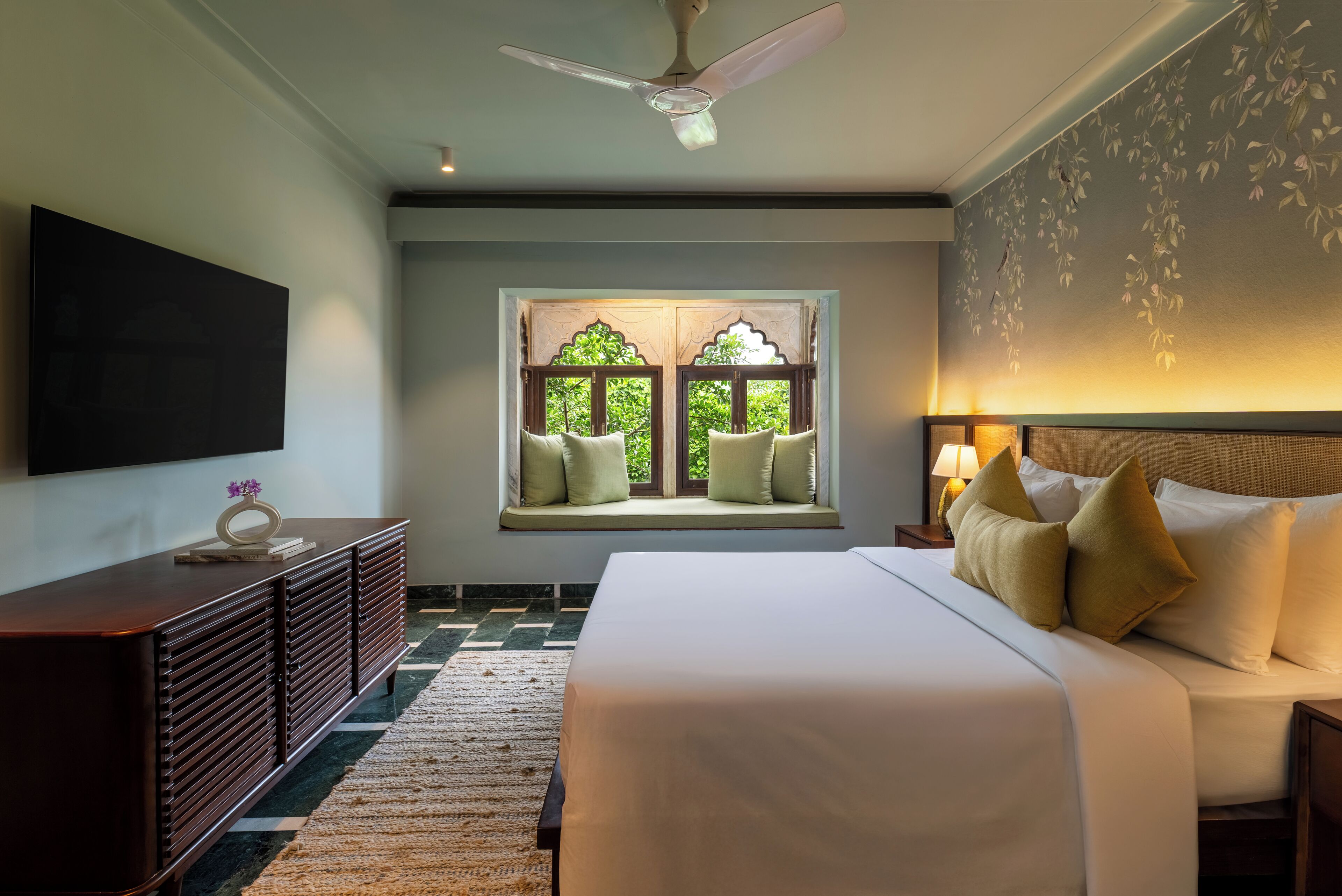 A hotel room with a bed, a nightstand, a TV, a window with a view of greenery, and a ceiling fan.