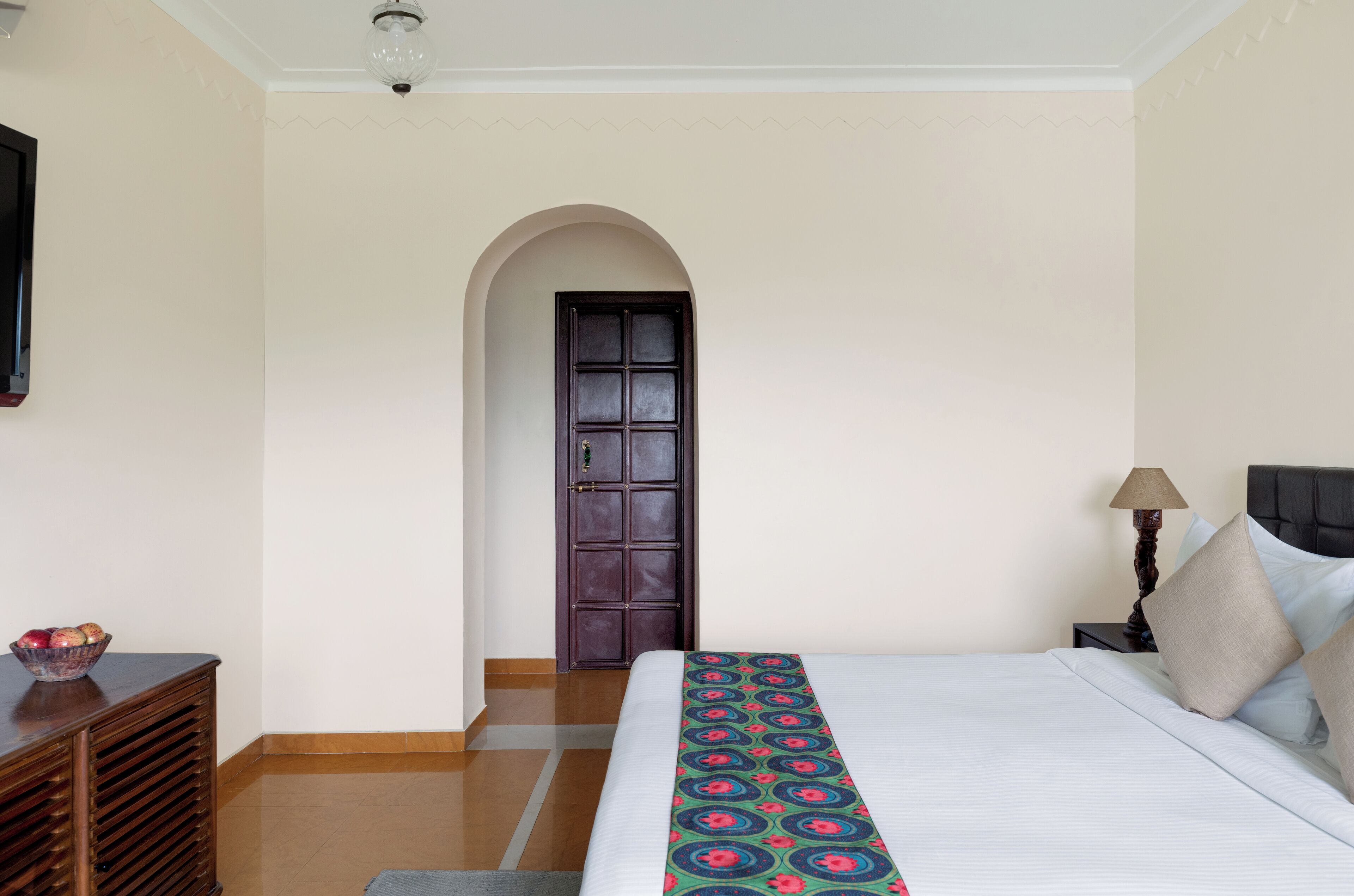 A hotel room with a bed, a nightstand, a lamp, a bowl of fruit, and a door.