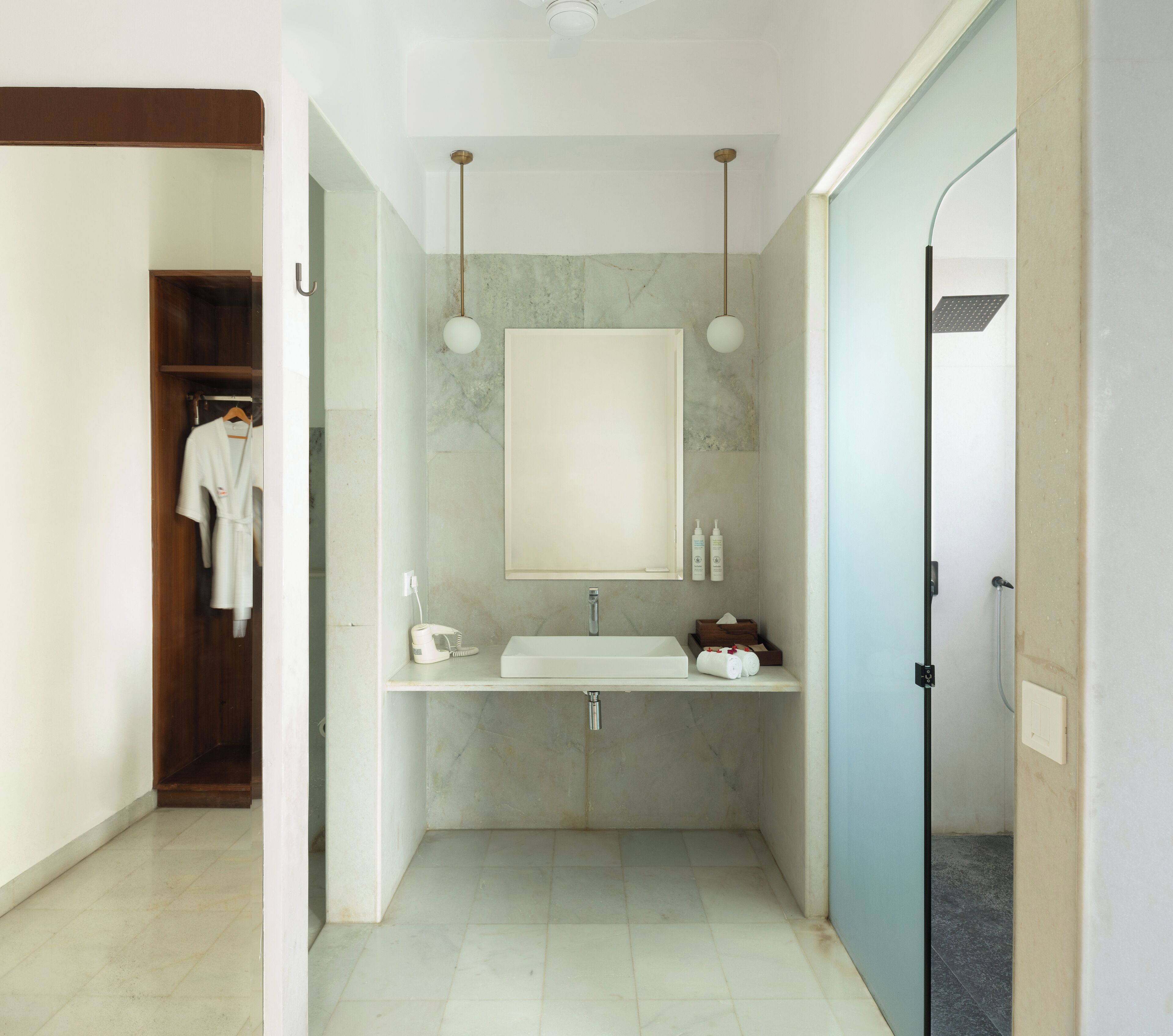 A modern bathroom with a large mirror, a sink, and a shower. There is a robe hanging on a door, and toiletries are placed on the sink counter.