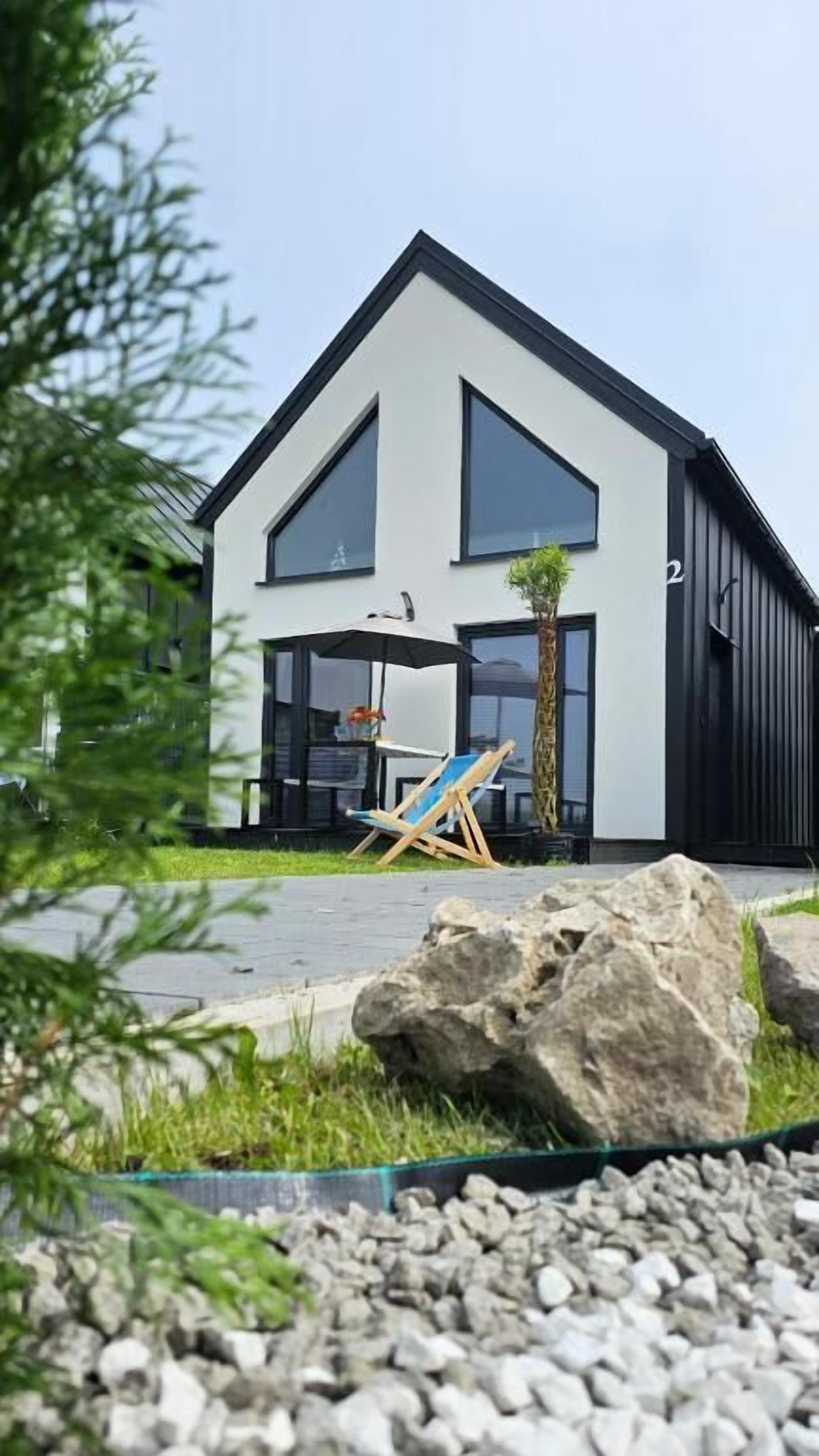 A modern two-story house with a large rock in the foreground, a chair, and a small palm tree.