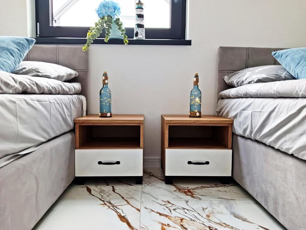 Quarto com duas camas, cada uma acompanhada de uma mesinha de cabeceira e uma gaveta. Há uma janela acima das camas com vista para um prédio e uma planta.