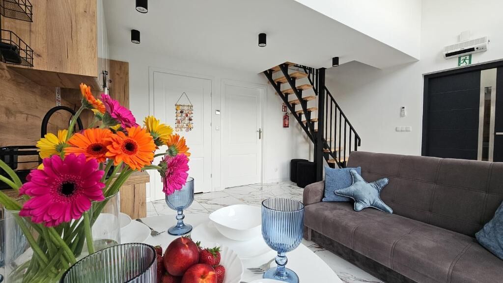 A modern living room with a brown sofa, a dining table with a vase of flowers, and a staircase in the background.