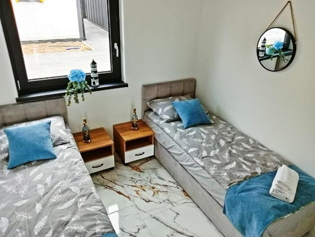 Une chambre avec deux lits simples, une table de chevet avec une lampe, une petite table ornée d’un objet décoratif et une fenêtre donnant sur l’extérieur.