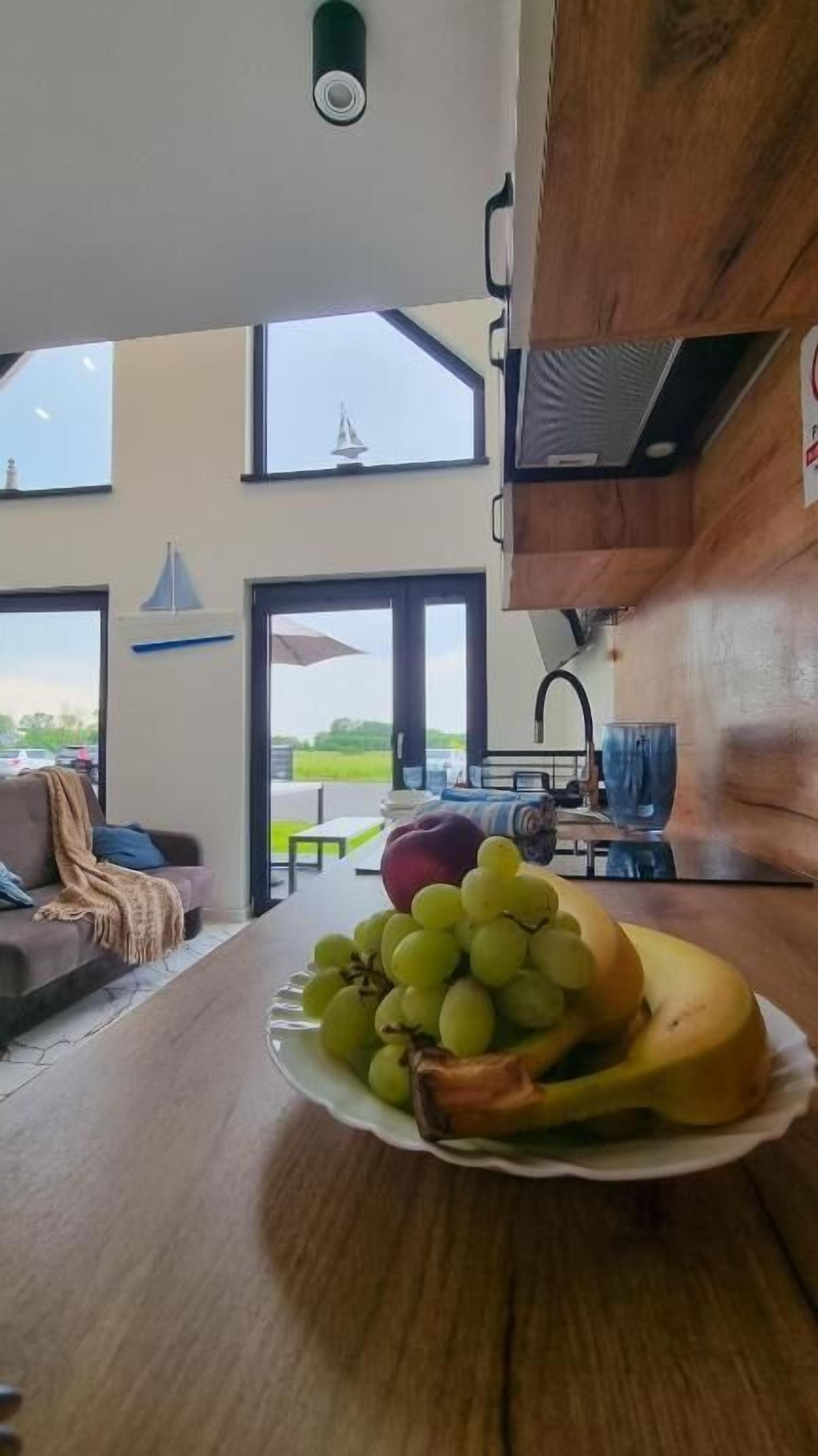 A kitchen with a large window, a dining table with a plate of fruit, and a view of a sailboat outside.