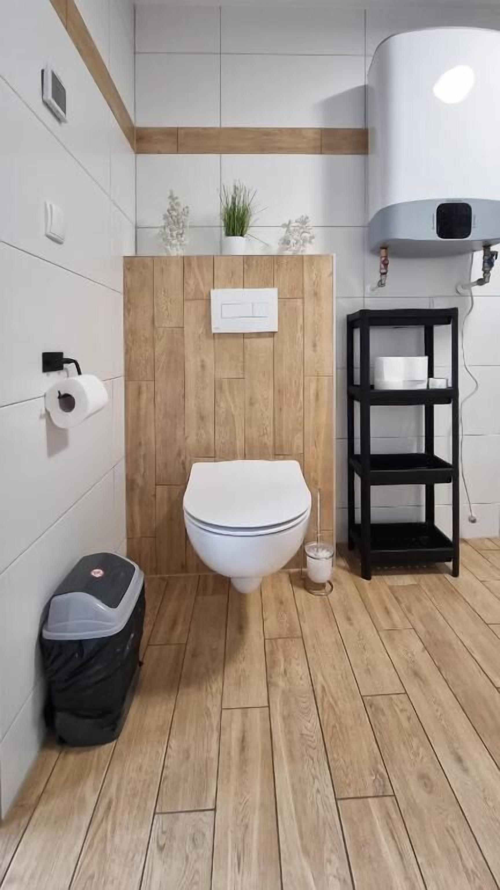 Une salle de bain moderne avec un plancher en bois, des murs blancs, des toilettes blanches, une étagère noire et un chauffe-eau blanc.