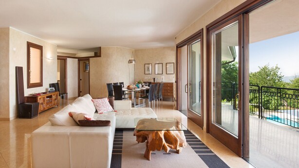 A spacious living room with a large sofa, a coffee table, and a wooden side table. There is a dining area with chairs and a table, and a balcony with a pool view.