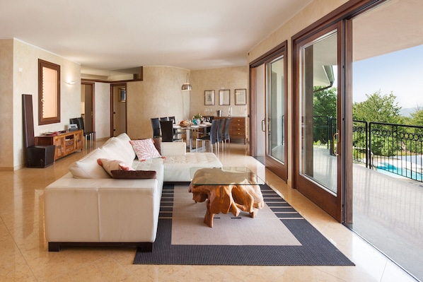 Uma sala de estar ampla com um sofá grande, uma mesa de centro e uma mesa lateral de madeira. Há uma área de jantar com cadeiras e uma mesa, e uma varanda com vista para a piscina.