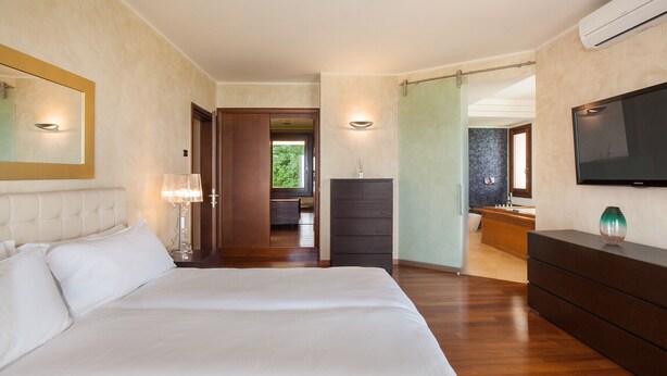 Ein Schlafzimmer mit einem großen Bett, einem Schminktisch, einem an der Wand montierten Fernseher und einem durchgehenden Türrahmen sichtbaren Badezimmer.