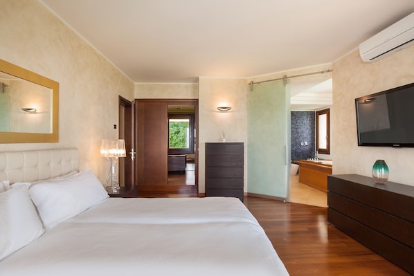 Een slaapkamer met een groot bed, een dressoir, een tv aan de muur en een zichtbare badkamer door een geopende deur.