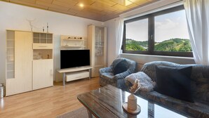 A living room with a TV, sofa, and a large window with a view. - Apartment 'Wohung Zum Ausblick' with Mountain View, Balcony and Wi-Fi (Plettenberg)