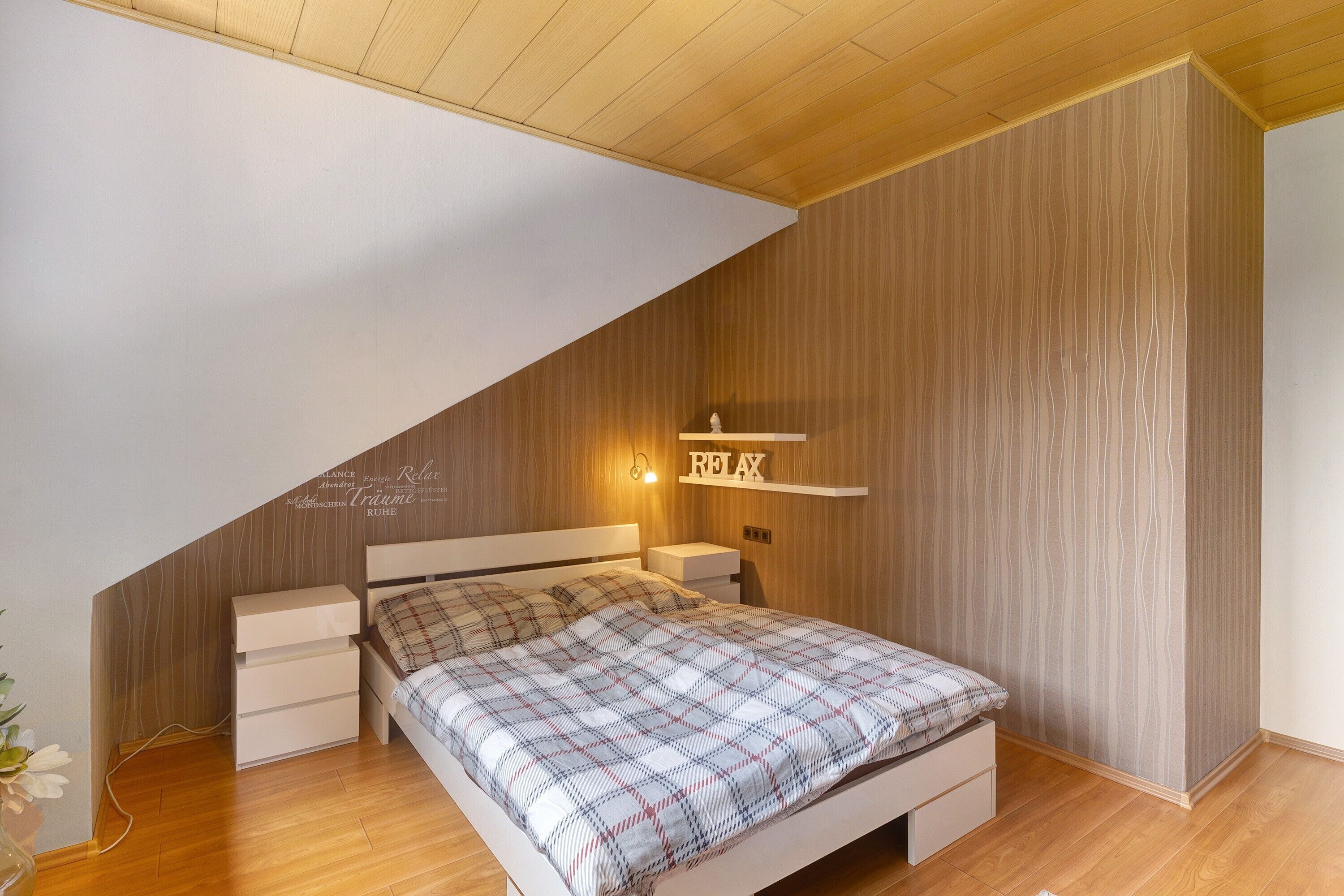 A bedroom with a sloped ceiling, a bed with a plaid bedspread, bedside tables, and a wall-mounted shelf with a sign.