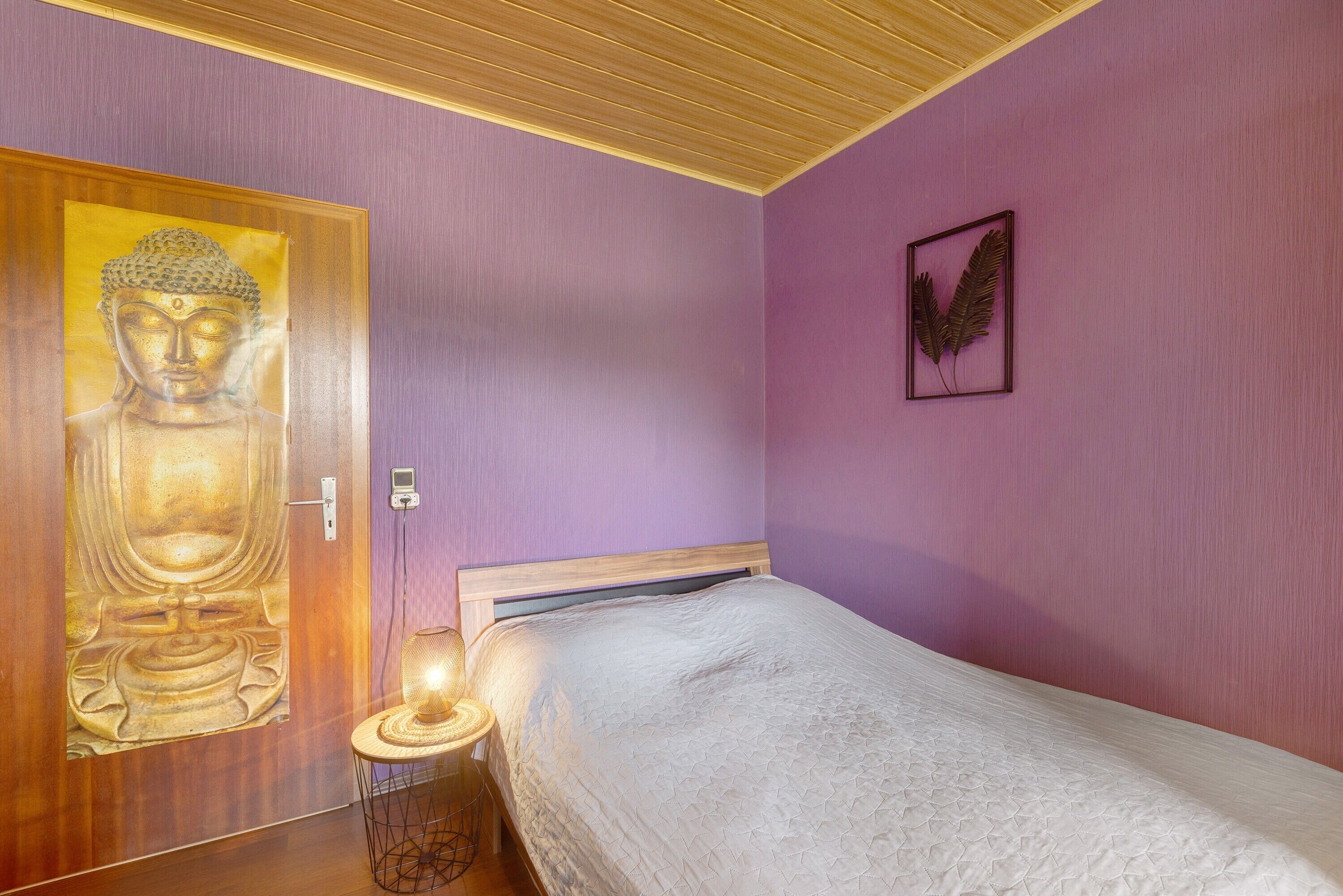 Ein Zimmer mit einem Bett, einer Holztür, einer Buddha-Statue, einer Wandpflanze und einem Nachttisch mit einer Lampe.