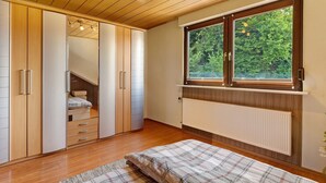 A bedroom with a wooden ceiling, a large window with a view of greenery, a bed with a plaid bedspread, and a wardrobe with sliding doors. - Apartment 'Wohung Zum Ausblick' with Mountain View, Balcony and Wi-Fi (Plettenberg)