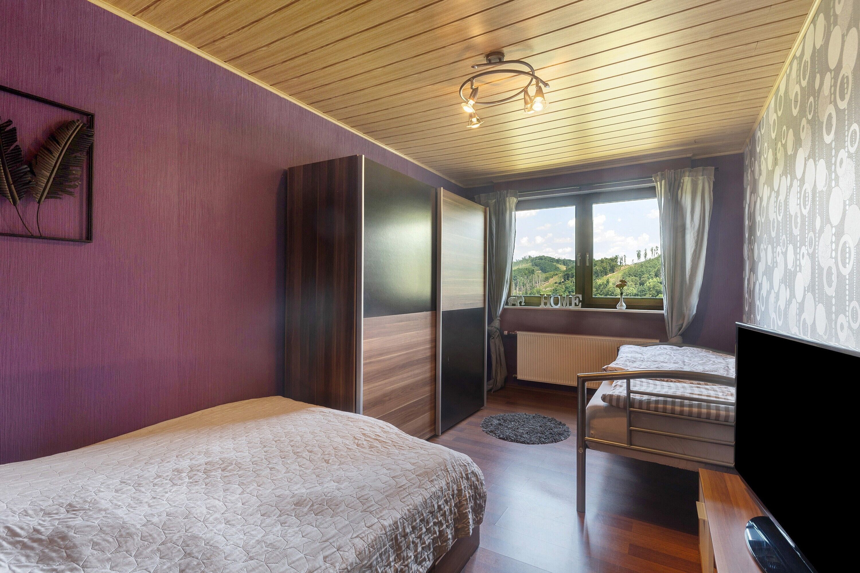 A bedroom with two beds, a desk, a chair, a TV, a window with curtains, and a view of greenery outside.