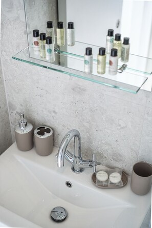 A bathroom sink with a chrome faucet, a glass shelf above holding various toiletries, and a beige mug on the sink.