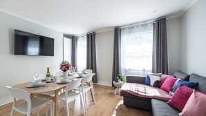 Una sala de estar moderna con comedor, cortinas grises, un televisor de pantalla plana y un sofá gris con cojines de colores.