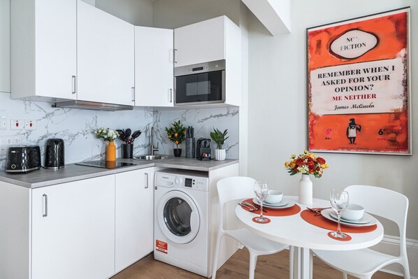 Une cuisine moderne avec des meubles blancs, un four encastré et un lave-linge. On y trouve un coin repas avec une table et des chaises blanches, ainsi qu’une affiche accrochée au mur.
