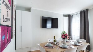 A dining area with a round table set for four, white chairs, a flat-screen TV mounted on the wall, and a window with curtains.