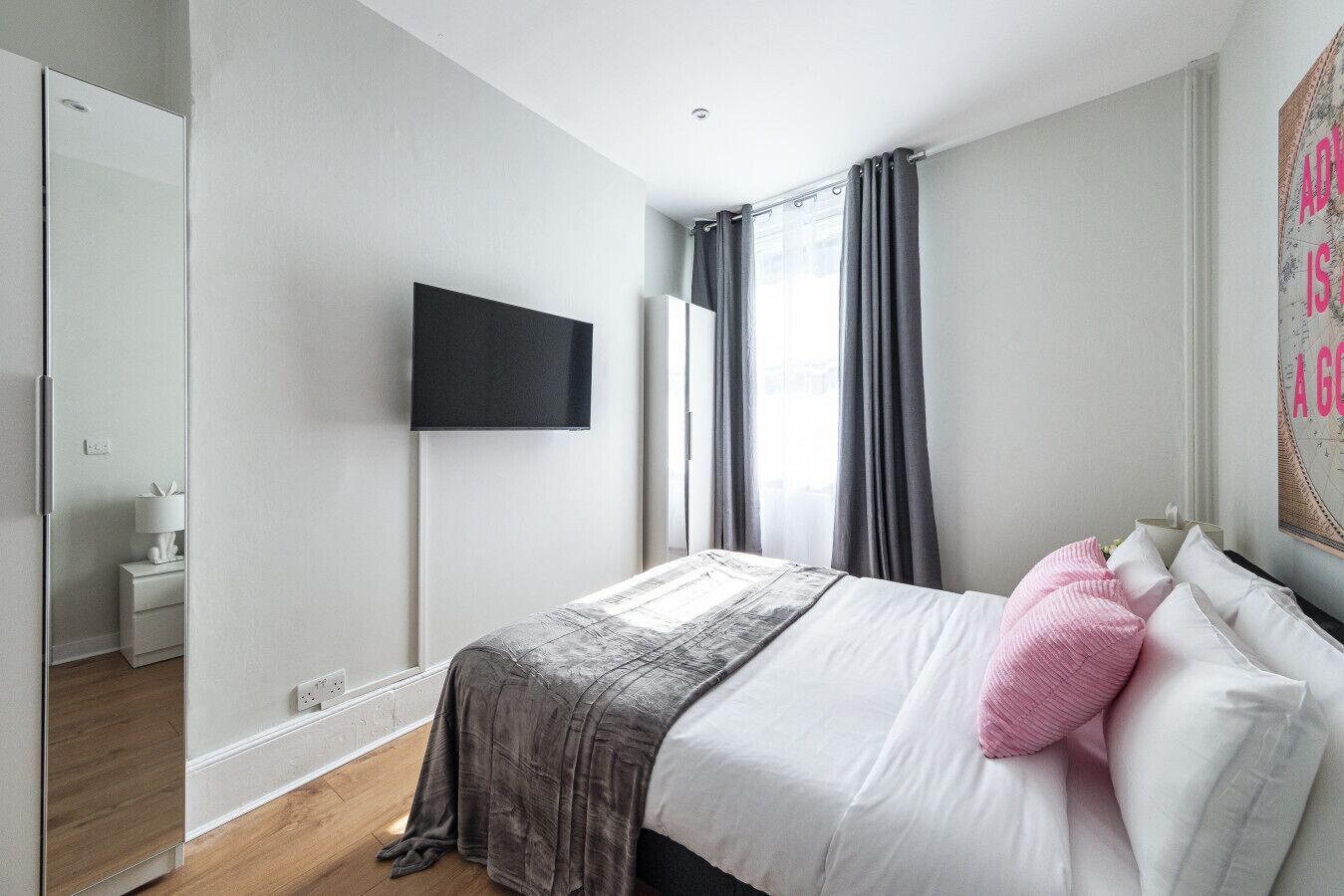 Um quarto moderno com cama, televisão de tela plana, janela com cortinas e espelho fixado na parede.
