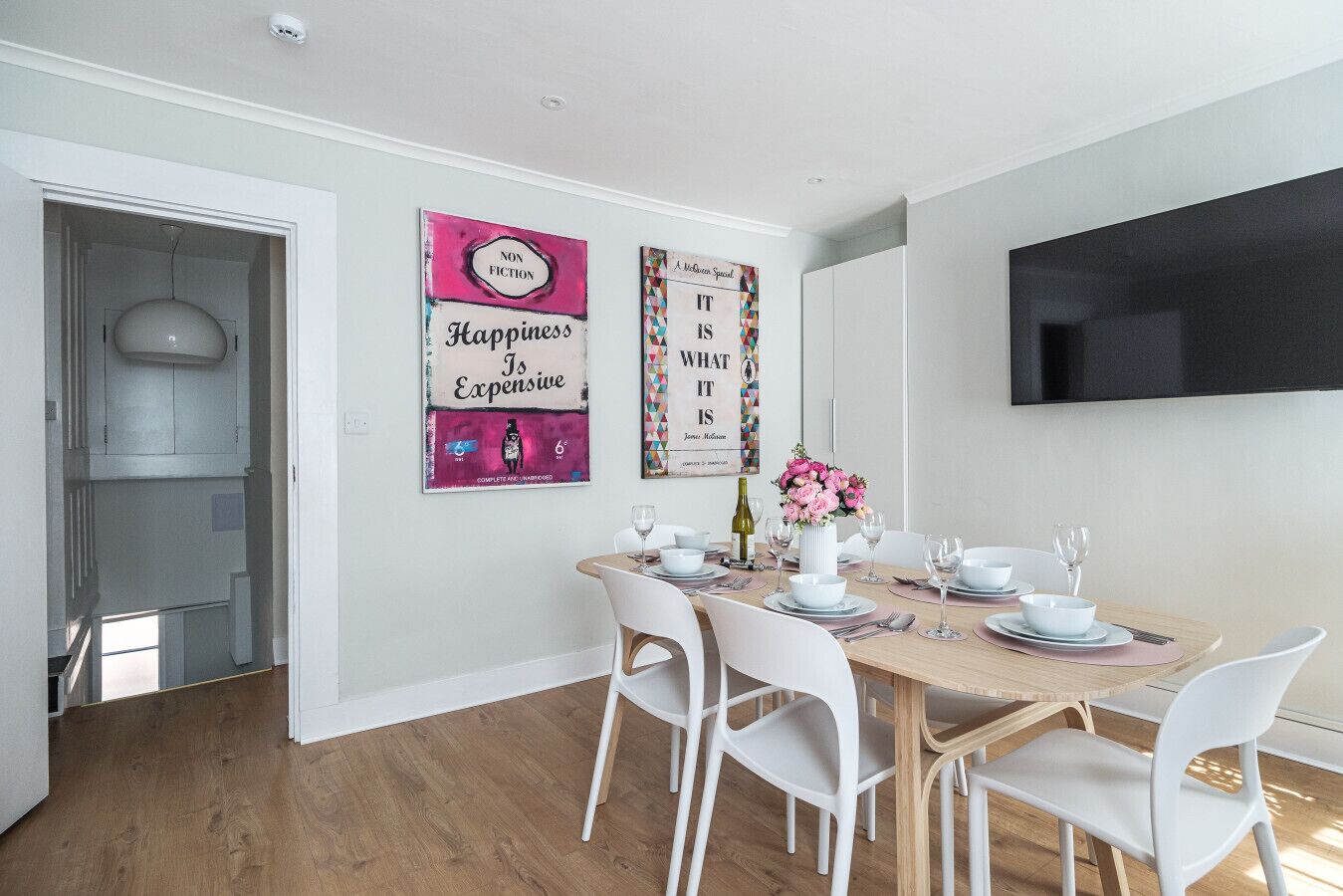 Een eetgedeelte met een tafel voor vier personen, witte stoelen, een bloevas en twee kunstwerken aan de muur.