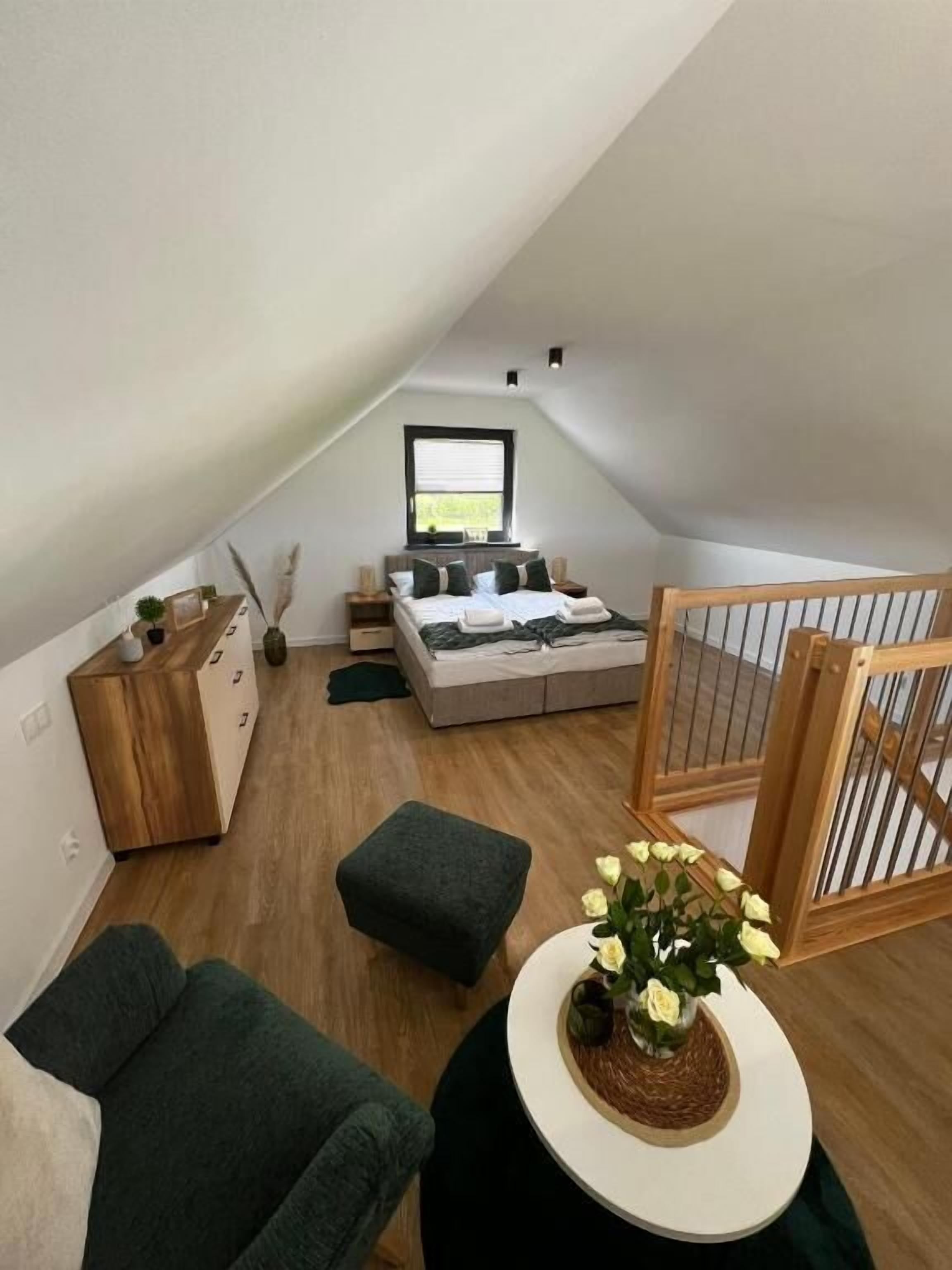 A modern loft bedroom with a sloped ceiling, a bed with a view of the outdoors, a wooden dresser, a dark green sofa, and a small round table with flowers.