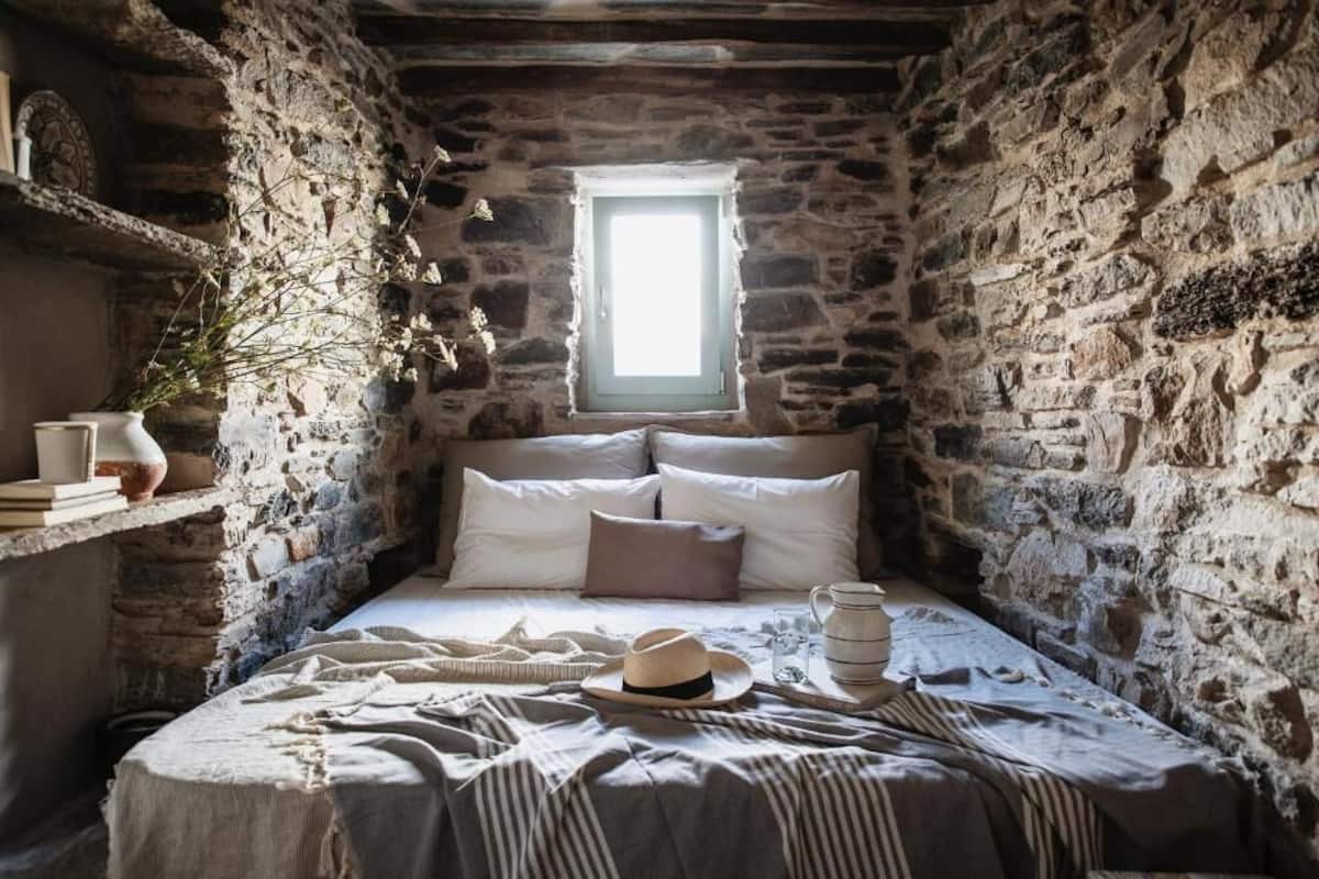 A narrow stone-walled room with a bed, a window, and a shelf with books and a vase.