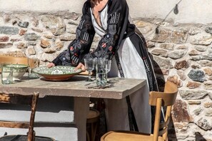 A person seated at a table with a stone wall background, a wooden chair, and a table with a bowl and glasses. - Onos Eco Living (Tinos)