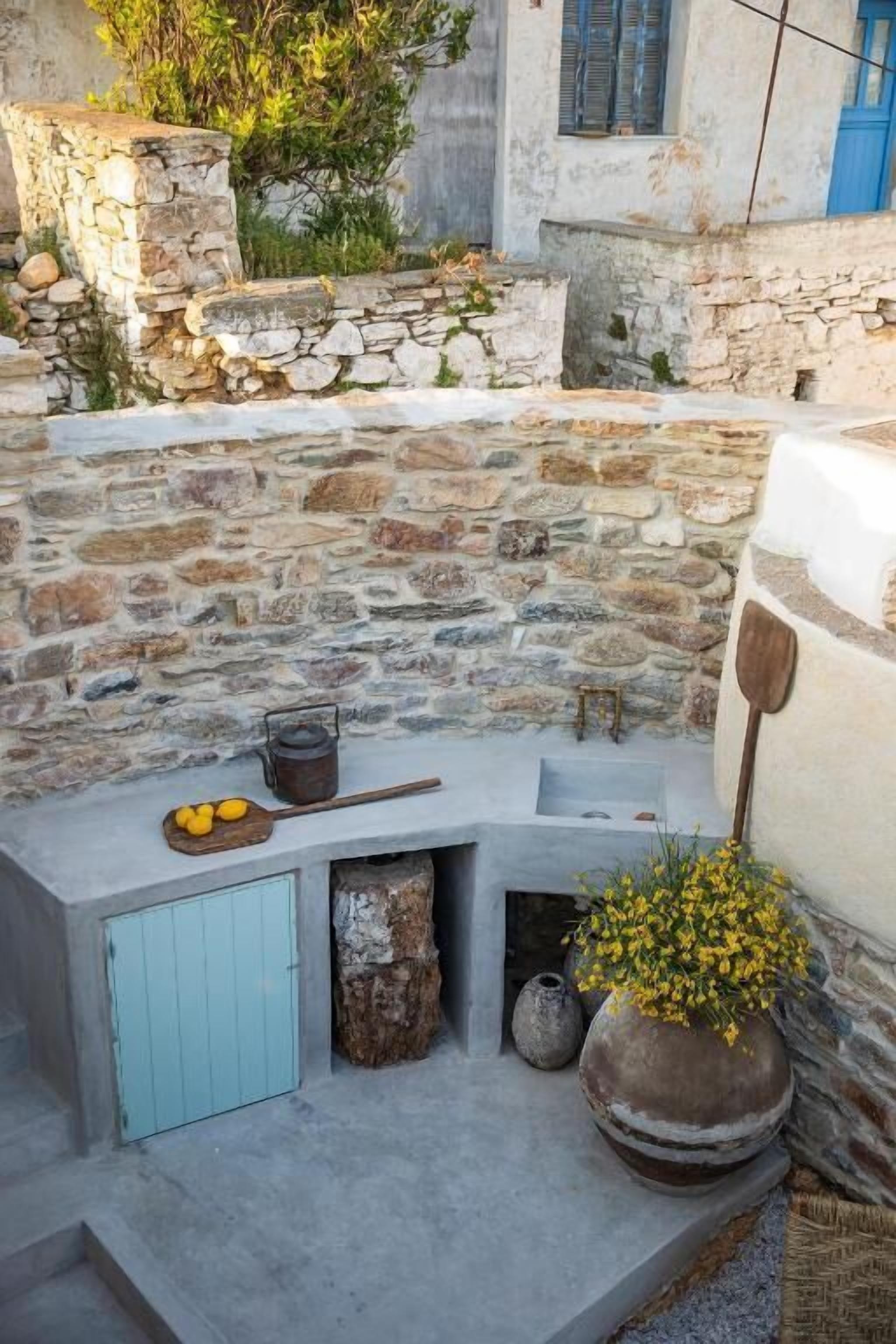 Un espace extérieur entouré de murs en pierre, avec un banc en pierre rustique, une petite table ornée de citrons, une cuillère en bois et une plante en pot.