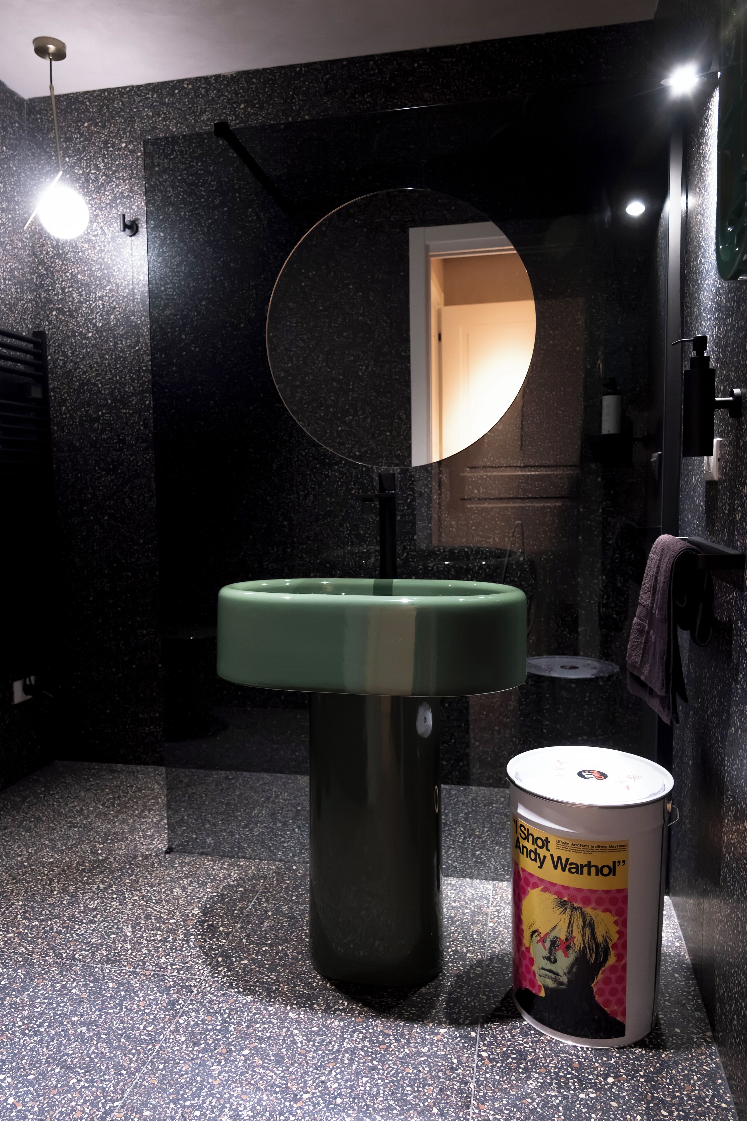 Un baño con un lavamanos verde, un espejo redondo y un cubo de basura con una impresión de Andy Warhol.