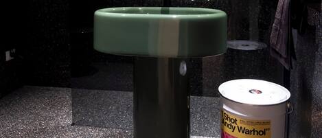 Un baño con un lavamanos verde, un espejo redondo y un cubo de basura con una impresión de Andy Warhol.