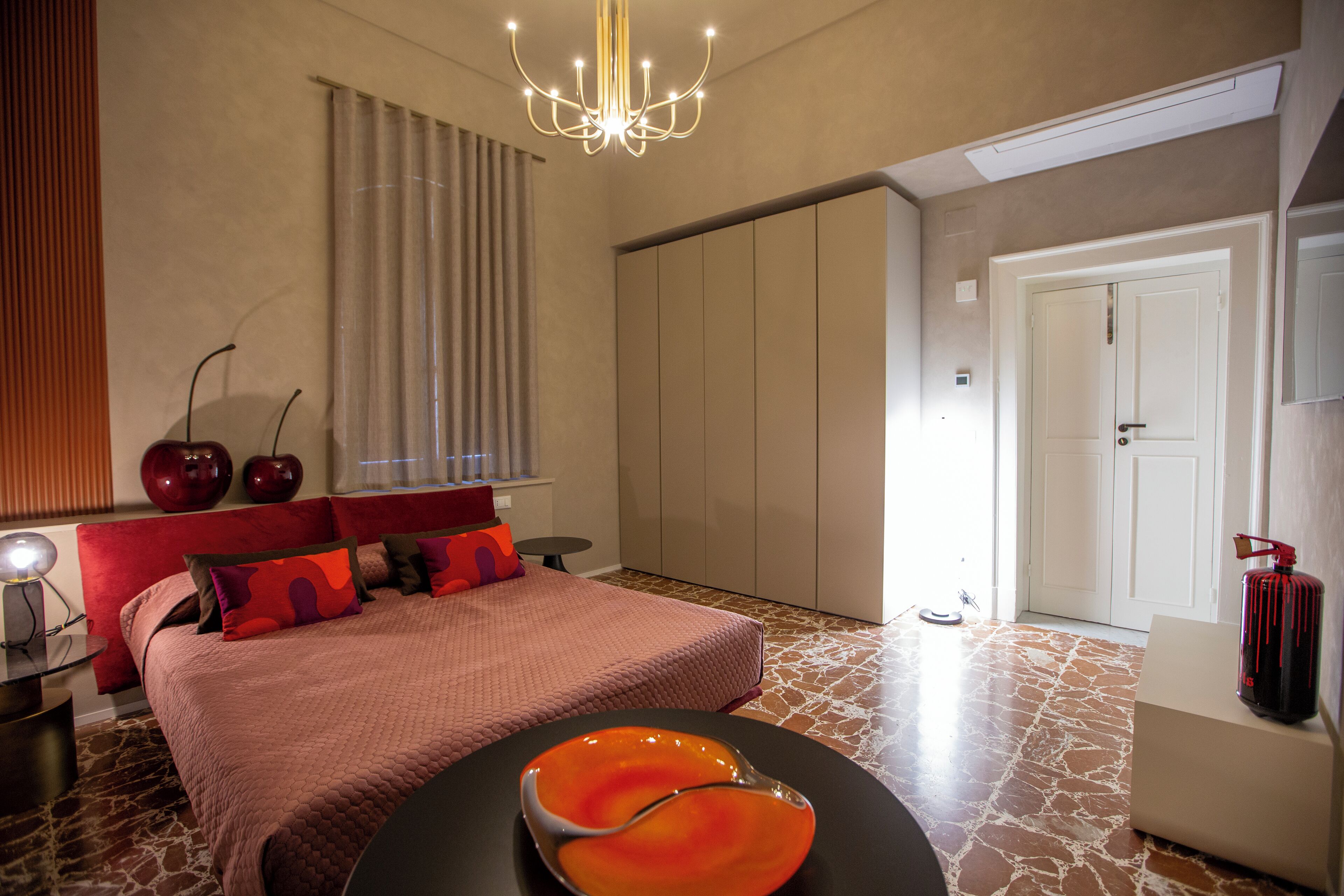 Een moderne slaapkamer met een groot bed, een kroonluchter en een opvallende oranje schaal op de bijzettafel.