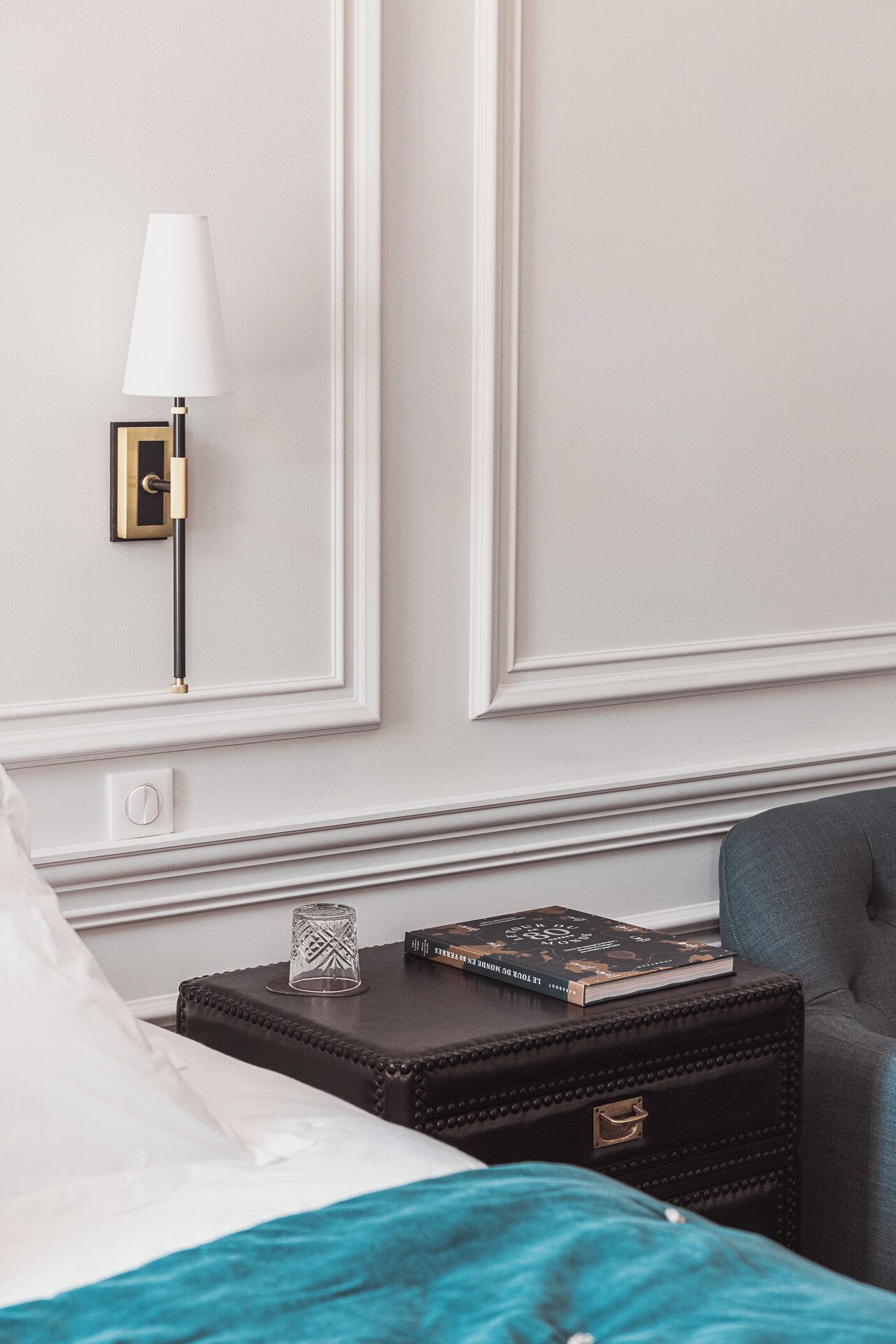 Una cama bien hecha con un edredón color turquesa, una mesita de noche con un libro y un vaso, y una lámpara fijada a la pared.