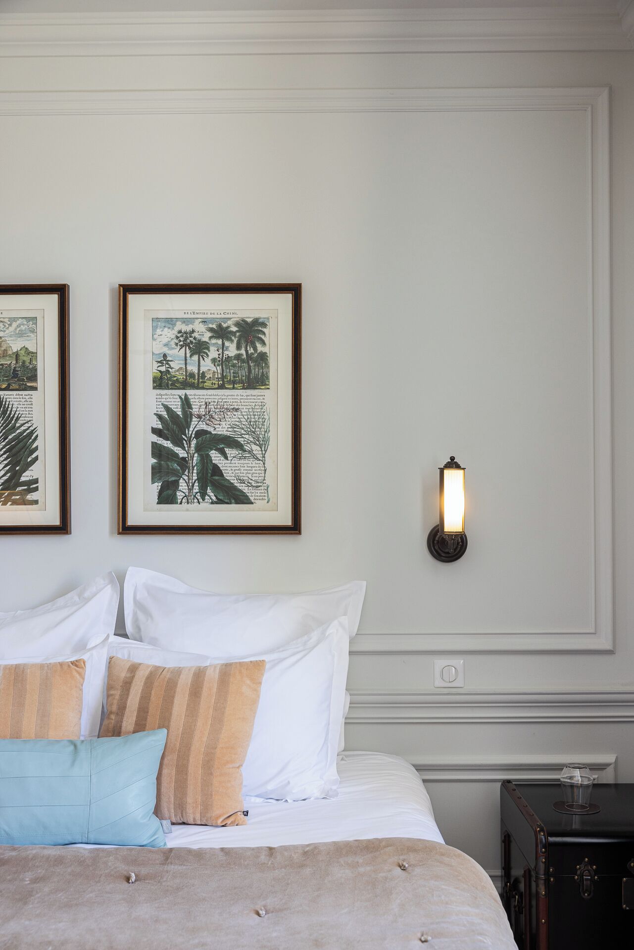 A neatly made bed with white and striped pillows, a framed botanical illustration, and a wall-mounted light fixture.