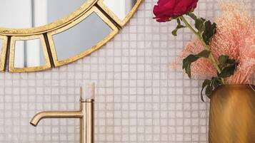 Un baño con lavamanos blanco, grifo dorado y espejo con marco dorado. En la encimera hay un florero con flores rosas y una caja dorada.