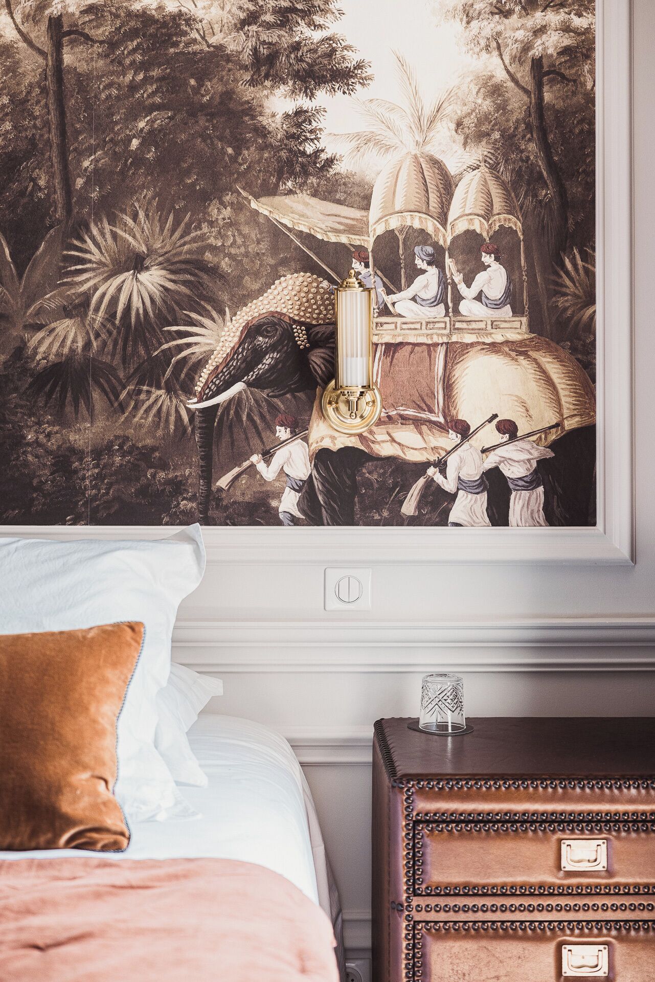A bedroom with a large wall mural, a bed with white and brown pillows, and a wooden chest of drawers.