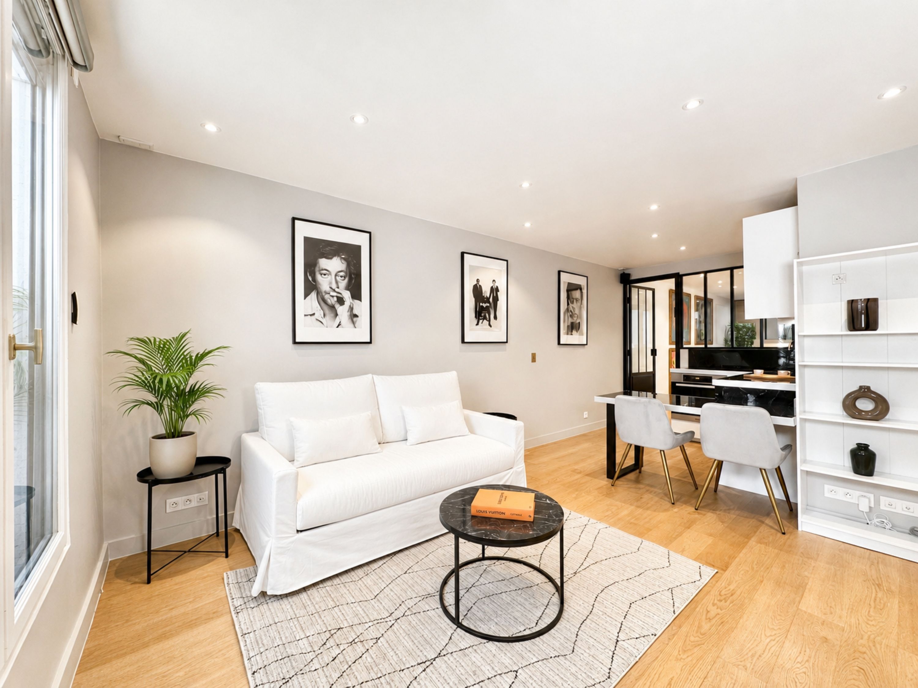 Una sala de estar moderna con un sofá blanco, una mesa de centro y una planta en maceta. Hay cuadros enmarcados en la pared y un comedor con mesa y sillas.