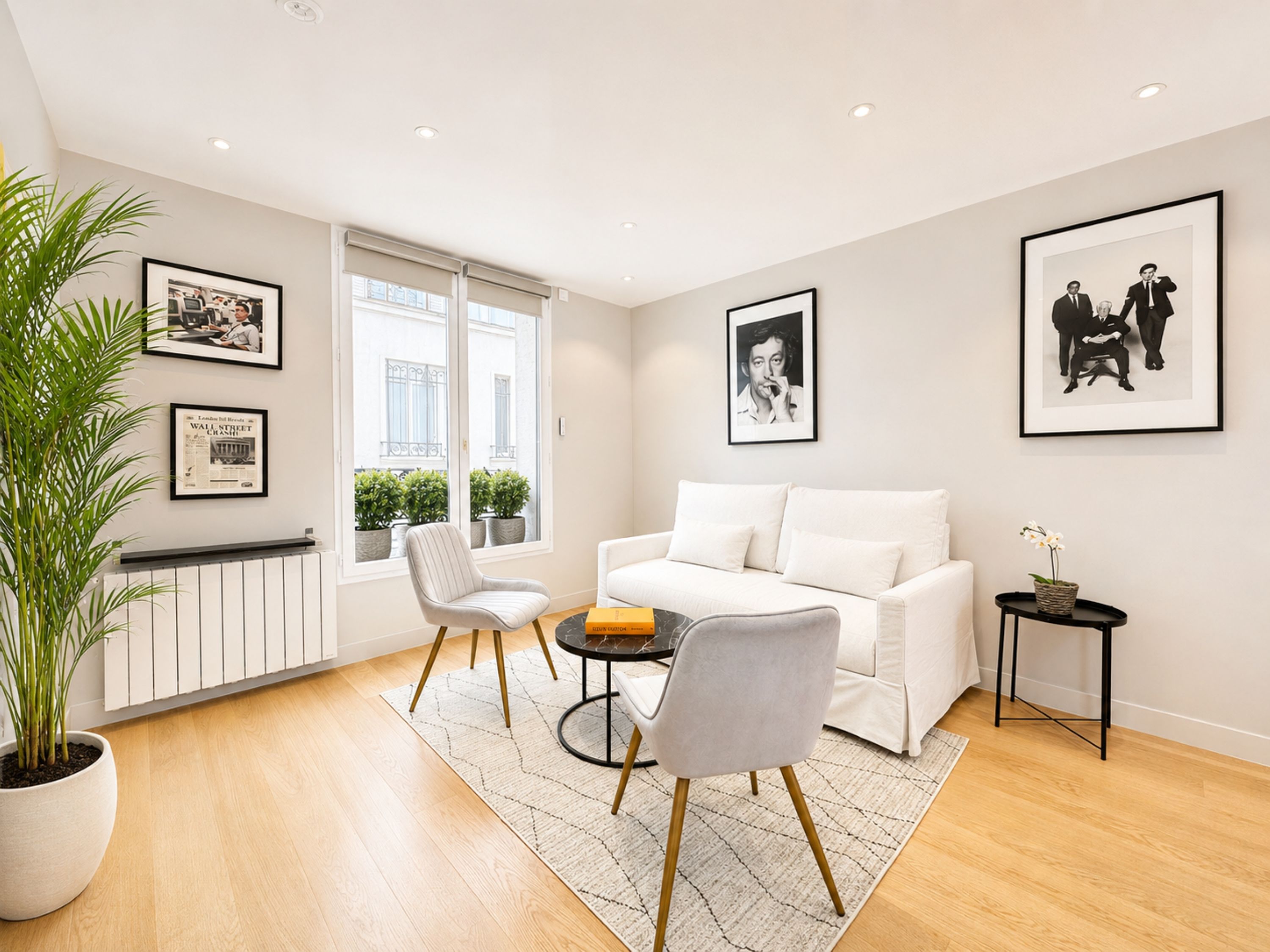 Una sala de estar moderna con un sofá blanco, dos sillones, una mesa de centro y una planta en maceta. Hay cuadros enmarcados en las paredes y una ventana con vistas a edificios.