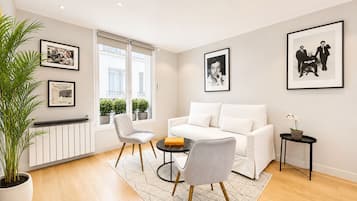 Una sala de estar moderna con un sofá blanco, dos sillones, una mesa de centro y una planta en maceta. Hay cuadros enmarcados en las paredes y una ventana con vistas a edificios.