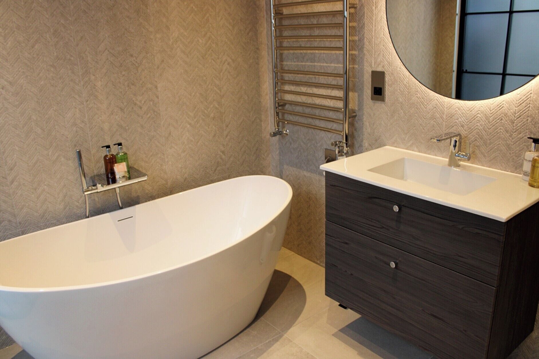 Ein modernes Badezimmer mit einer freistehenden Badewanne, einem Waschtisch mit Spiegel und Pflegeprodukten auf einem Regal.