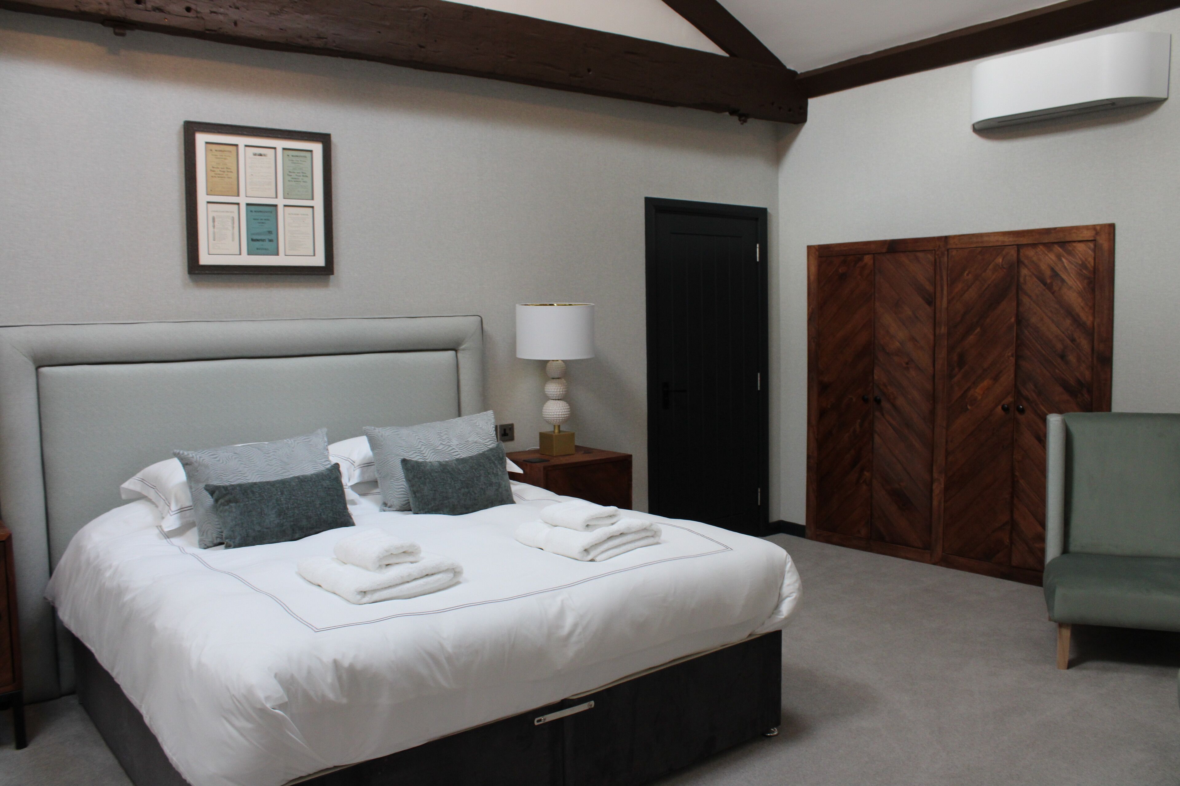 Una cama bien hecha con almohadas y toallas dobladas, una lámpara de noche, un armario de madera y un sillón verde en una habitación de diseño sencillo y limpio.