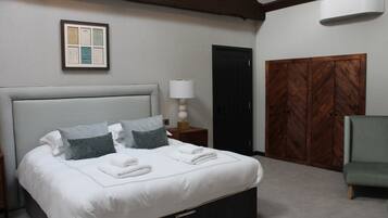 Una cama bien hecha con almohadas y toallas dobladas, una lámpara de noche, un armario de madera y un sillón verde en una habitación de diseño sencillo y limpio.