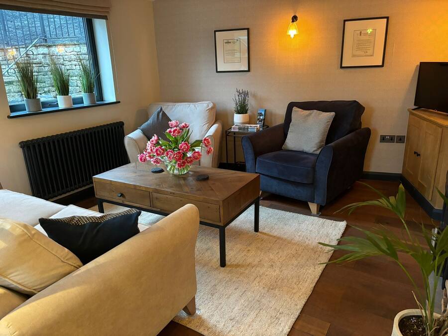Uma sala de estar aconchegante com sofá, poltrona e uma mesa de centro com um vaso de flores. Há uma janela com plantas no parapeito, um radiador e um abajur fixado na parede.
