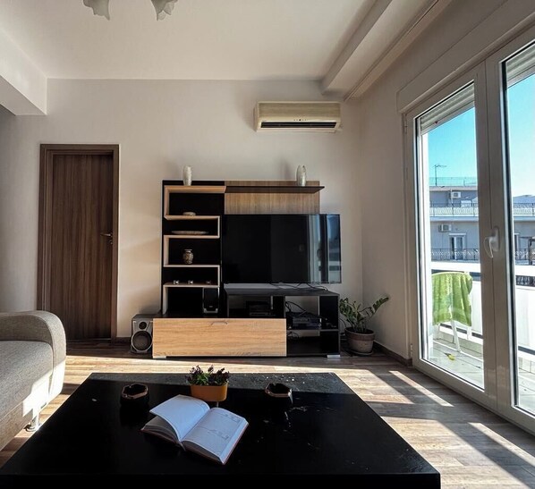 A modern living room with a large flat-screen TV, wooden entertainment unit, and a sliding glass door leading to a balcony.