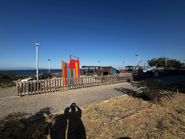 Un parque infantil en la playa, con valla de madera, estructura de juego de colores y vista despejada al mar.