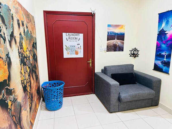 A laundry room with a red door, a blue laundry basket, and a grey sofa. - Su Casa Hostel (Riyadh)