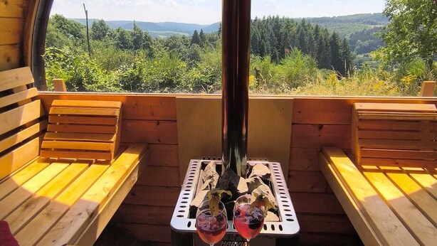 A cozy wooden cabin with a circular window offering a scenic view of a forested landscape. Inside, there is a wooden bench and a table with two glasses of red wine. - Nature & wellness ''on the Hardt'' *Jacuzzi *Sauna *all in the forest (Mürlenbach)