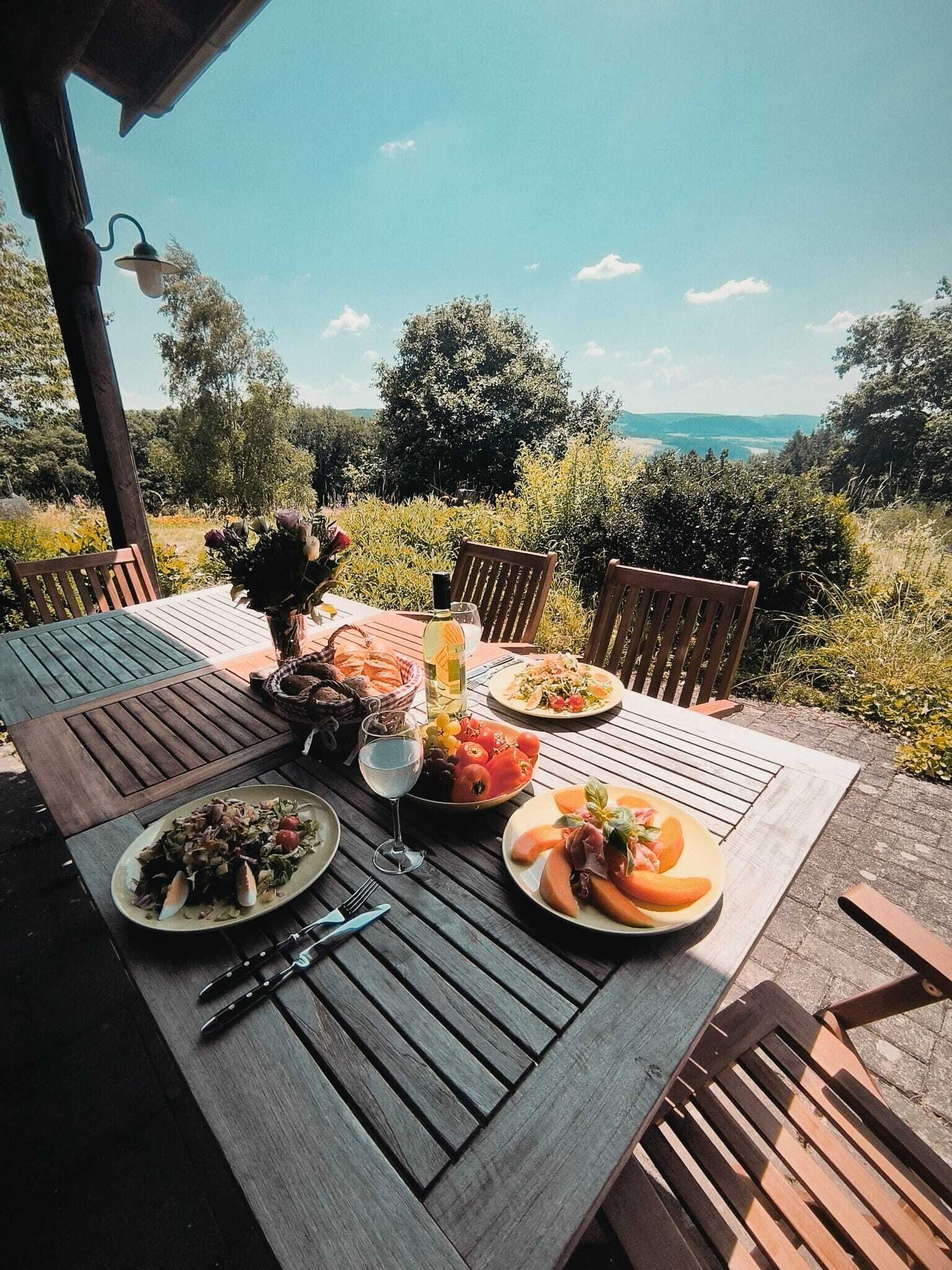 Una mesa de comedor al aire libre con varios platos, una botella y un vaso de agua, con un paisaje de árboles y colinas de fondo.