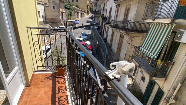 Una calle urbana estrecha flanqueada por edificios, un balcón con tejas y una planta en maceta, y una pequeña estatua.