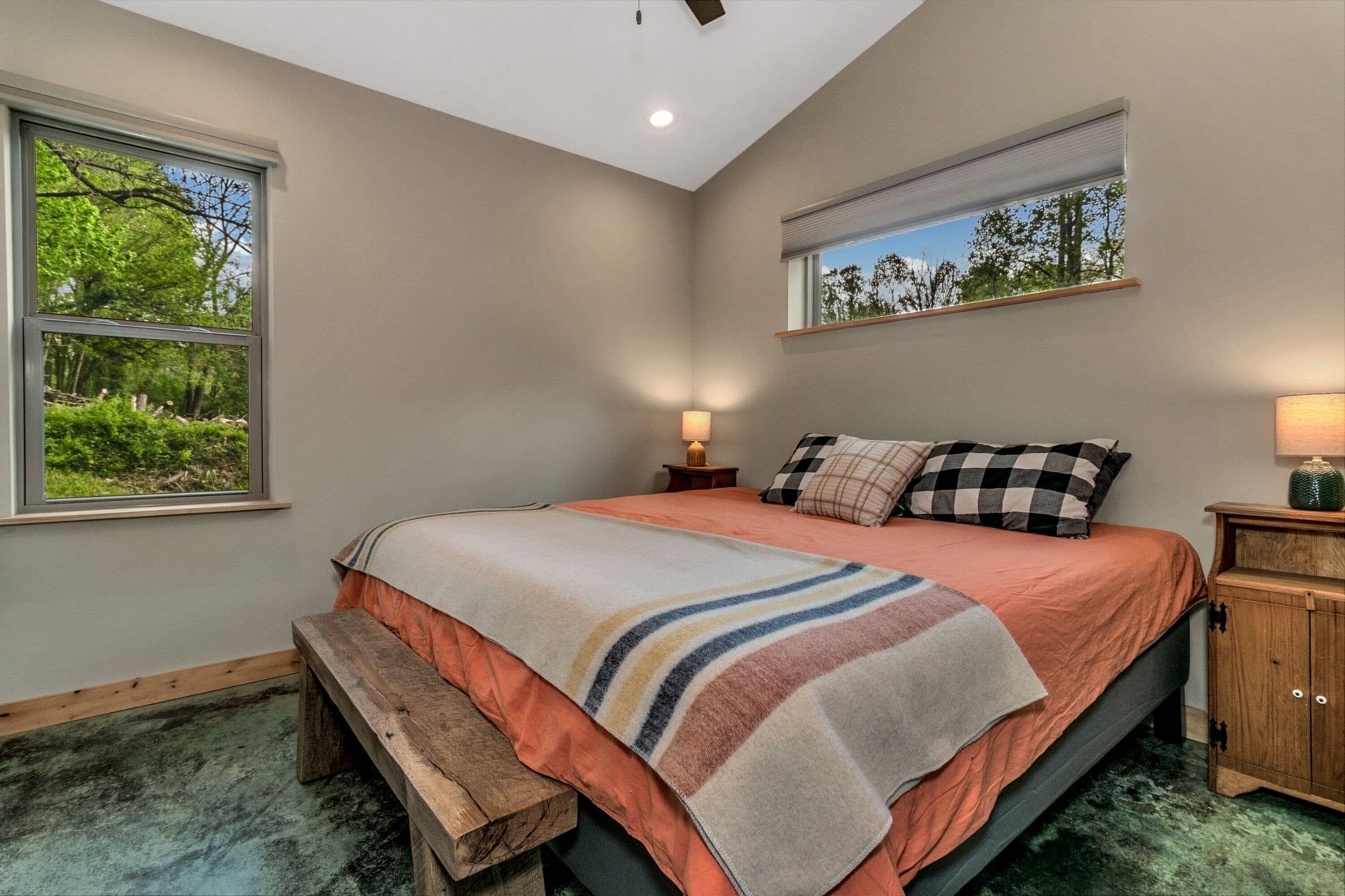A bedroom with a large bed, a bench, a nightstand, and a window with a view of greenery.