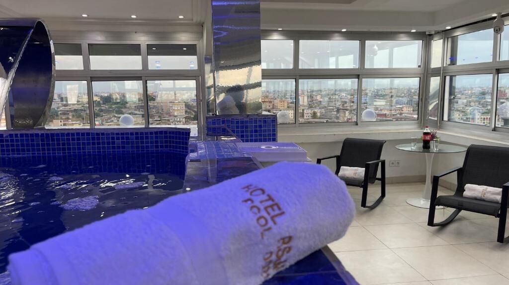 Habitación de hotel con un ventanal amplio, vista a la ciudad, una piscina y una mesa con sillas.